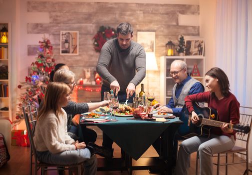 Cheerful girl singing on guitar while his father is slicing the chicken at christmas family celebration.