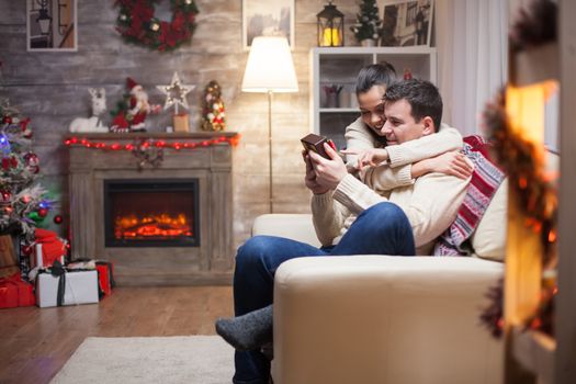 Happy young man with his gift from wife on christmas day sitting on couch with fireplace in living room.