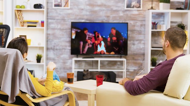 Young couple sitting comfortable on in front of tv eating chips and popcorn looking at their cat.