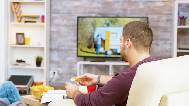 Rear view of young man sitting on comfortable chair eating fried chicken with his girlfriend in front of tv.