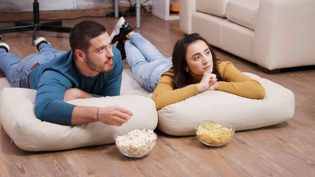 Young man and woman sitting on the floor eating chips and popcorn while watching tv.