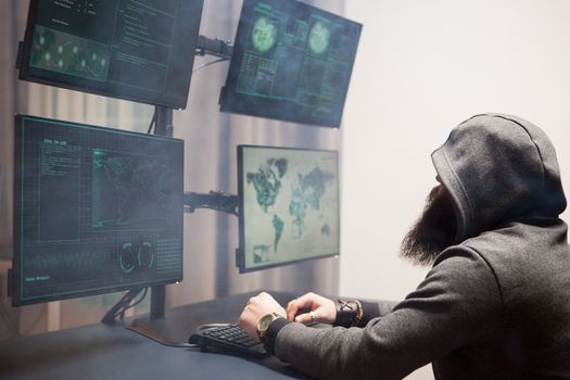Side view of bearded hacker with a hoodie using multiple screens for cyberterrorism.