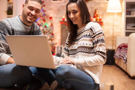 Happy couple having fun on internet using laptop on christmas day in front of warm fireplace.