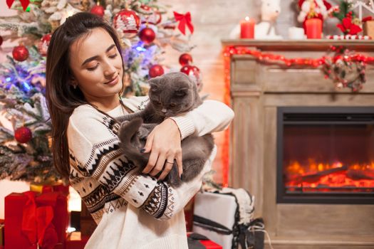 Cheerful young woman looking at her cat in front of fireplace celebrating christmas.