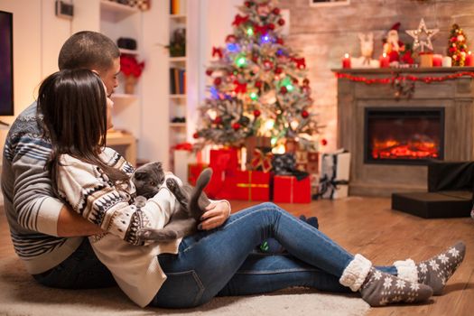 Couple in matching clothes and their scottish fold sitting together on the floor in front of fireplace celebrating christmas.