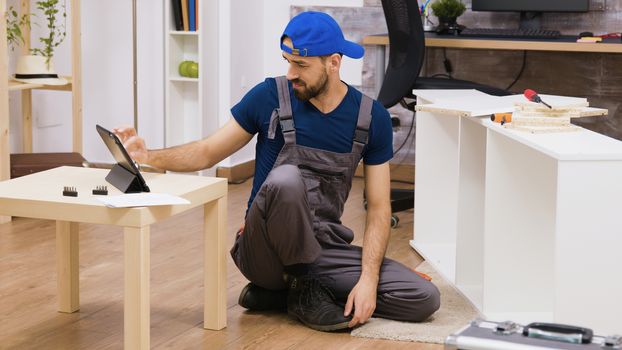 Male worker in overalls assembles new white furniture in new home owners. Man reading instruction on laptop.