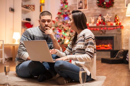 Happy couple thinking about the gifts they have to buy for christmas using their laptop in front of fireplace. Glass of champagne.