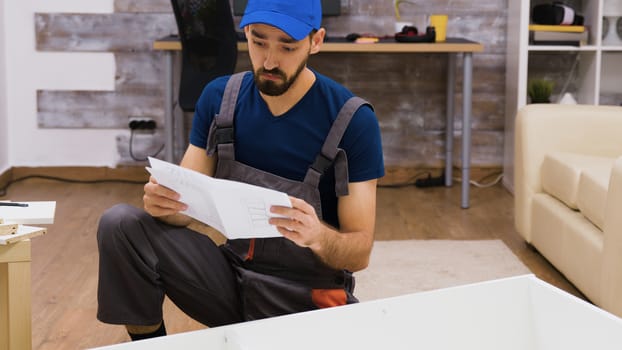 Confused worker in overalls reading instructions for furniture assembly wearing a cap.