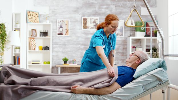 Female nurse helping an old disabled sick man who lies in hospital bed in a nursing retirement home