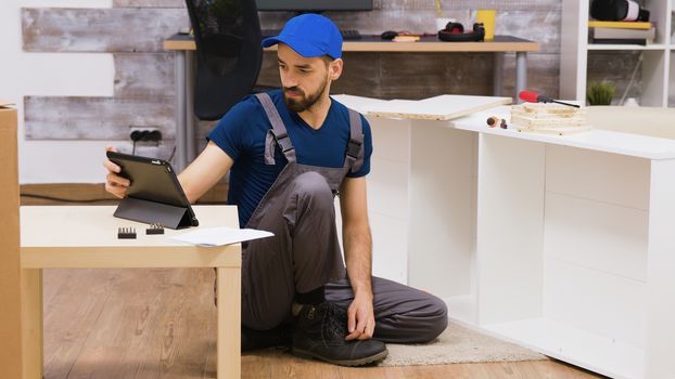 Male worker in overalls with a cap assembly a shelf in new home following insctruction from tablet computer