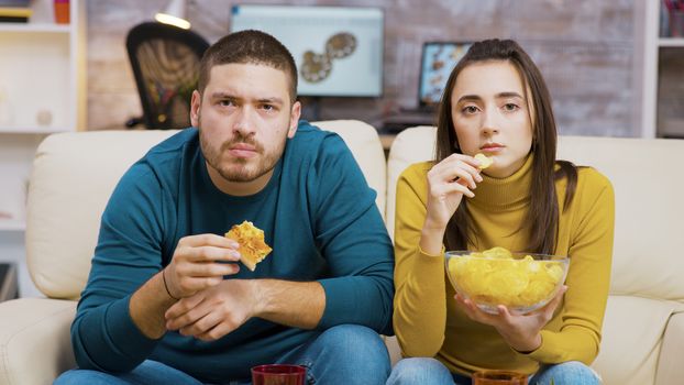 Scared couple while watching tv and eating junk food sitting on the couch.