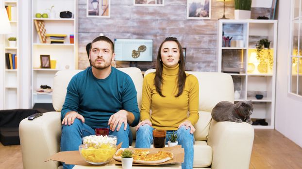 Disappointed couple after their favorite football team lost the game. Cat laying on sofa. Popcorn, pizza and soda on coffee table.