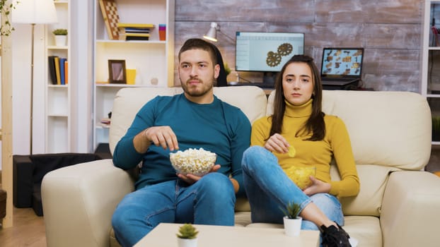 Beautiful young girl laying on her boyfriend's shoulder while watching tv and eating chips. Boyfriend eating popcorn.