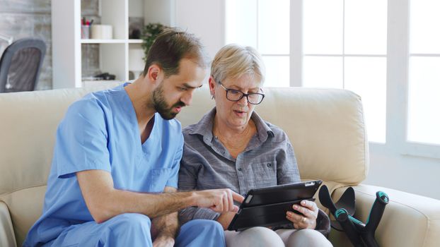 Male assistant teaching senior retired elderly old woman how to use her tablet computer in nursing home for her therapy