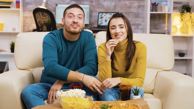Couple sitting on couch laughing while watching tv and eating pizza. Popcorn and pizza on coffee table.
