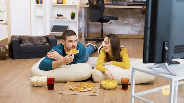 Bearded man drinking soda and sitting on pillows for the floor with his attractive girlfriend watching tv while the cat is sleeping in the background.