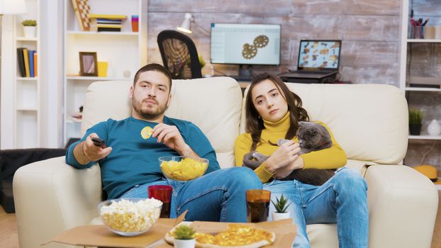 Young couple relaxing on the couch with their cat watching tv. Man eating chips.