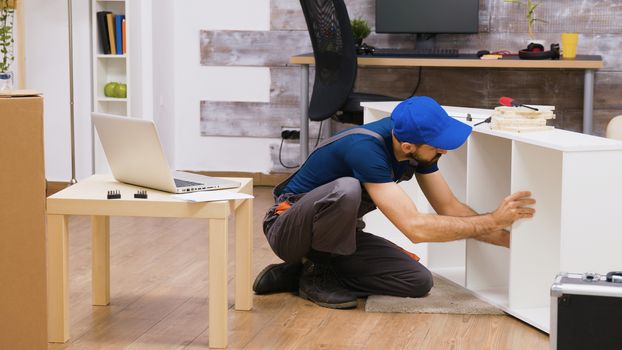 Professional worker in overalls consults furniture assembly instructions from laptop. Worker wearing a cap.