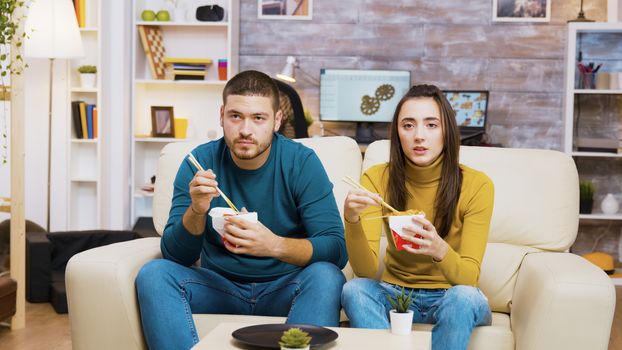 Couple sitting on couch eating noodles with chopstick and watching tv.