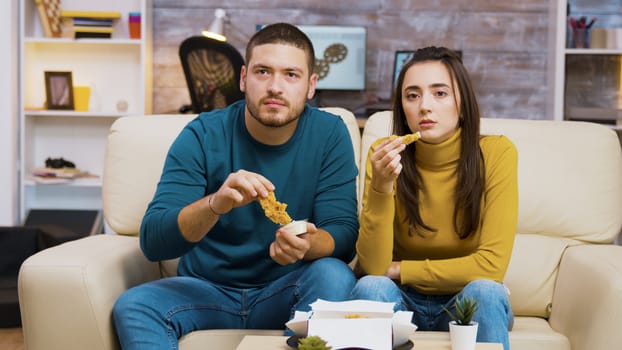 Young couple sitting on couch eating fried chicken while watching tv.