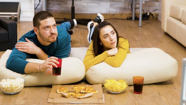Attractive couple relaxing on pillows for the floor watching tv and eating pizza with their cat sleeping in the background.