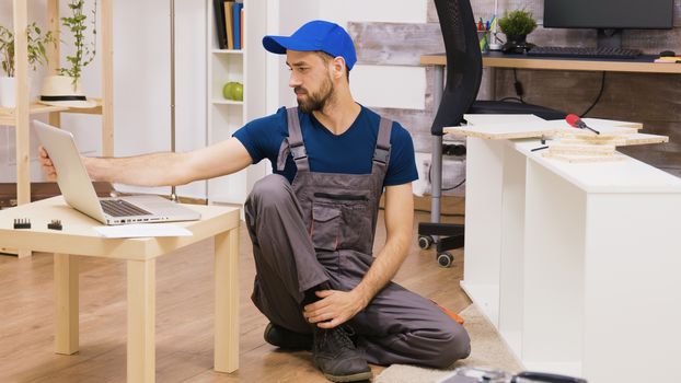 Professional worker assemble a shelf consults instructions on laptop. Handyman helping people who move in new home.