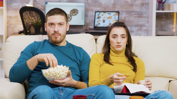 Scared couple watching tv eating pizza and popcorn sitting on couch. Couple eating junk food.