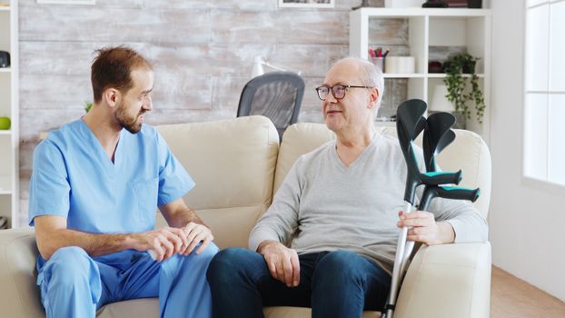 In retirement home male nurse is talking with an old disabled man with crutches next to him. The caregiver is consulting pensioner about drugs dosage