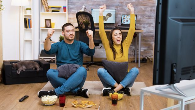 Caucasian young couple sitting on the floor in living room cheering up for their favorite sport team. Cat sleeping in the background.