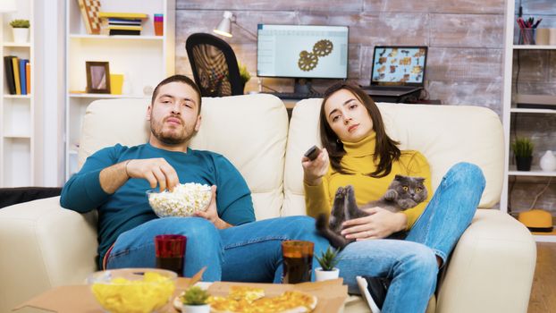 Bored girl sitting on the couch with the cat in her lap and boyfriend next to her is using tv remote control.