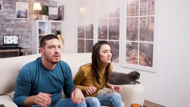 Upset young couple while watching a game on tv. Cat sitting on the couch. Pizza, soda, and popcorn on coffee table.