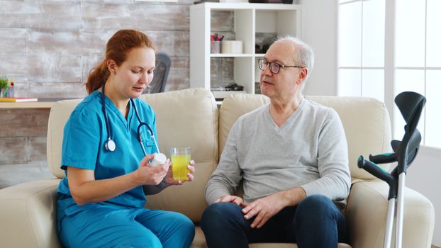 In retirement home nurse is giving daily pills to old man with crutches next to him. Caregiver and social worker