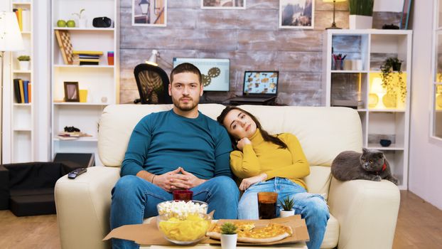 Bearded man changing tv channels with hand gestures while his girlfriend and the cat are next to him on the couch.