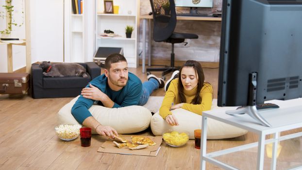 Bearded man drinking soda and sitting on pillows for the floor with his attractive girlfriend watching tv while the cat is sleeping in the background.