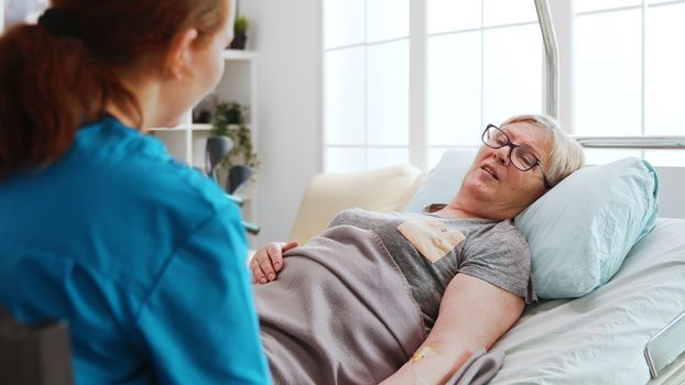 In nursing home old sick lady lying in hospital bed talks with a female nurse. Over the shoulder shot