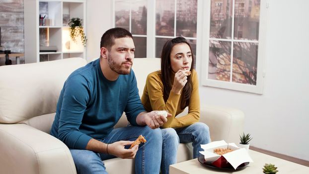 Concentrated young couple eating fried chicken while watching tv. Couple eating junk food.