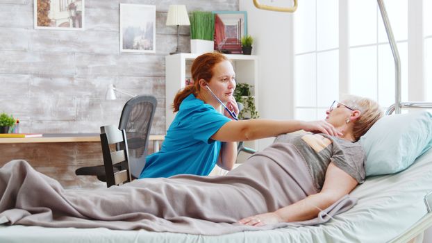 Female nurse listening to old lady heartbeats. She is lying in a hospital bed in a bright cozy nursing home