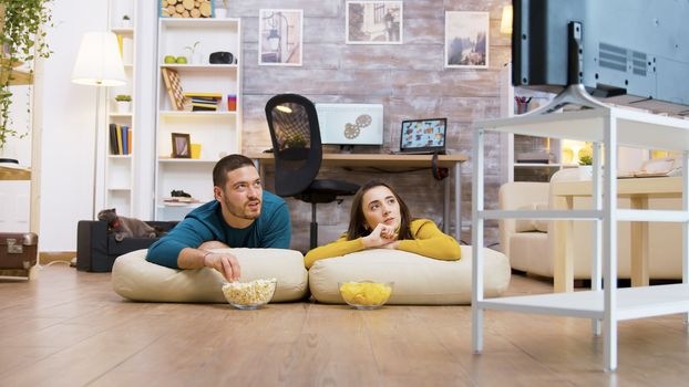 Couple talking with each other watching tv sitting on the pillows for the floor while their cat is relaxing in the background.