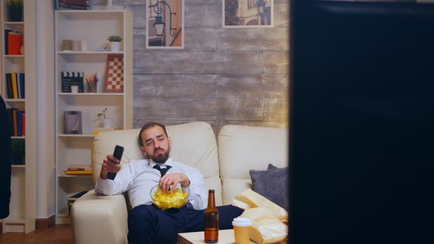 Tired and bored businessman with tie relaxing watching tv eating chips using remote control.