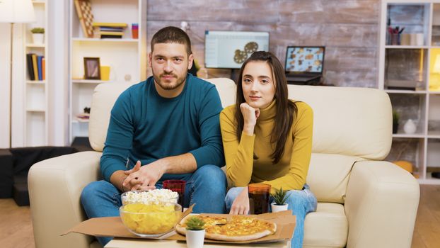 Couple sitting on couch laughing while watching tv and eating pizza. Popcorn and pizza on coffee table.