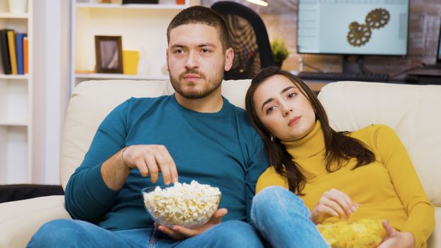 Beautiful young girl laying on her boyfriend's shoulder while watching tv and eating chips. Boyfriend eating popcorn.