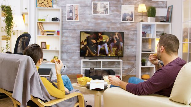 Back view of young couple eating fried chicken sitting comfortable in their chairs looking at tv. Cat relaxing on her cozy bed.