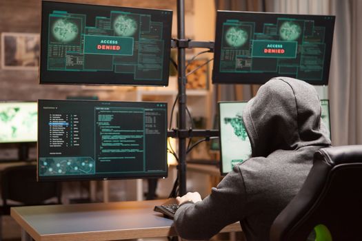 Back view of male hacker wearing hoodie while doing cyberterrorism using computer with multiple screens. Access denied.