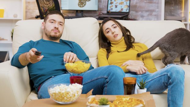 Young couple relaxing on the couch with their cat watching tv. Man eating chips.