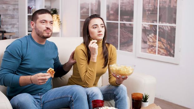 Scared couple after a frightening moment in the movie from tv. Couple sitting on couch eating pizza.