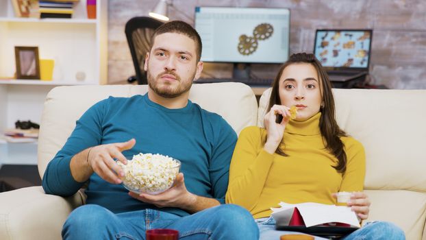 Scared couple watching tv eating pizza and popcorn sitting on couch. Couple eating junk food.