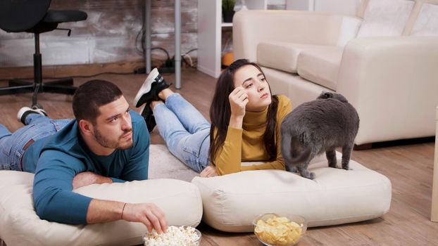 Young couple spending time with their cat while watching tv. Couple sitting on the floor and eating chips and popcorn.