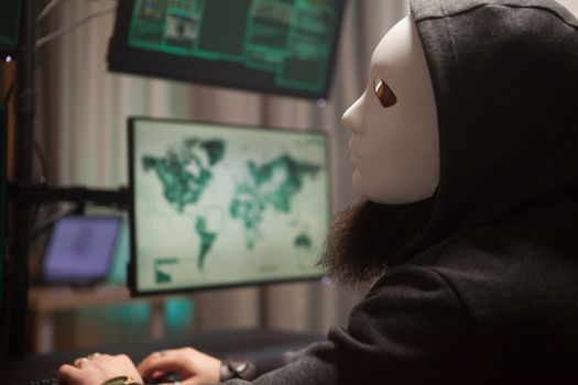 Bearded hacker with a hoodie wearing a white mask using his computer with multiple screens.