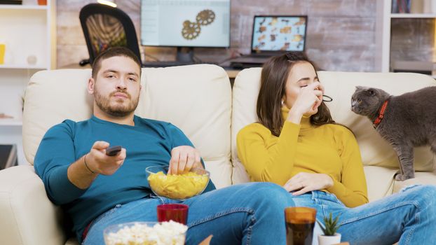 Bearded man eating chips and using remote control to change tv channels while his girlfriend is playing with the cat.