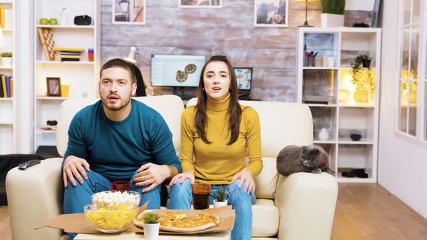 Disappointed couple after their favorite football team lost the game. Cat laying on sofa. Popcorn, pizza and soda on coffee table.